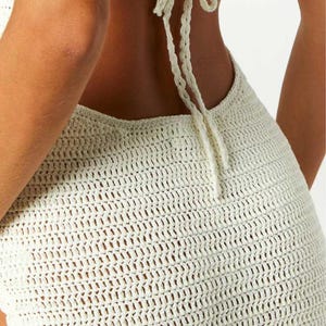 May include: Cream-colored crochet dress with a halter neck and open back. The dress features a textured, open weave design. The halter ties at the back of the neck. The dress is a summer or beachwear item.