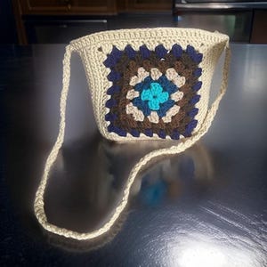 May include: A small crocheted bag with a brown, blue, and beige granny square design. The bag has a long strap and is made of a beige yarn.