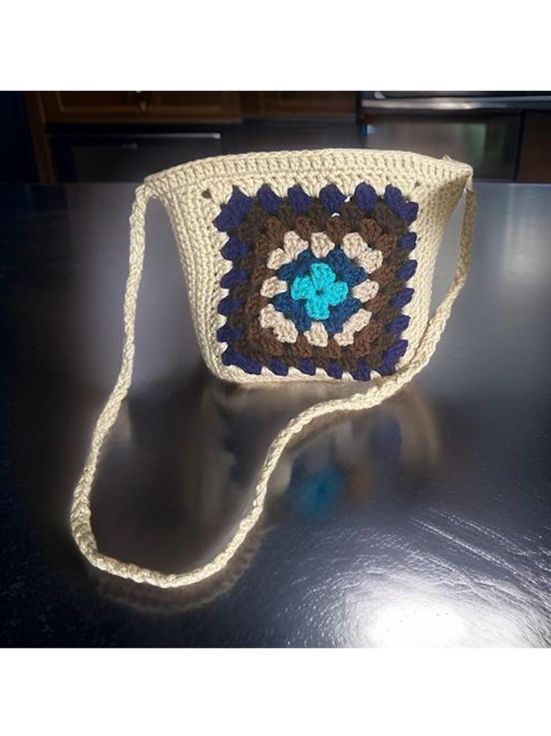 May include: A small crocheted bag with a brown, blue, and beige granny square design. The bag has a long strap and is made of a beige yarn.