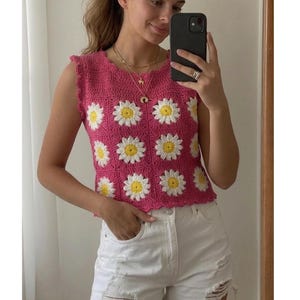 May include: A sleeveless, pink crochet top with a daisy pattern. The top features white and yellow daisy designs and a scalloped edge. The person in the image is wearing white denim shorts and holding a black smartphone.