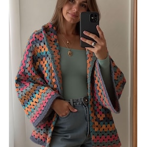 Może przedstawiać: Kolorowy, ręcznie robiony kardigan zrobiony na szydełku we wzór granny square. Kurtka ma mieszankę pastelowych kolorów, w tym różowy, pomarańczowy, niebieski i szary. Kardigan jest noszony na zielonym topie i dżinsowych szortach.