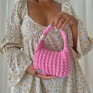 May include: A pink crocheted handbag with a small, rounded shape. The bag has a simple design with a single handle.