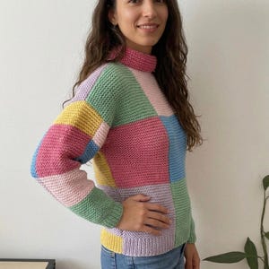 May include: A long-sleeve knit sweater featuring a patchwork design of large rectangular blocks in pastel pink, light blue, soft yellow, and mint green. The garment has a high mock neck collar and ribbed cuffs and hem in matching pastel colors.