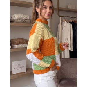 May include: A long-sleeve knit sweater featuring a color-blocked pattern of orange, pale yellow, white, and sage green squares. The sweater has a mock neck collar, ribbed cuffs, and a ribbed hem, paired with plain white pants.