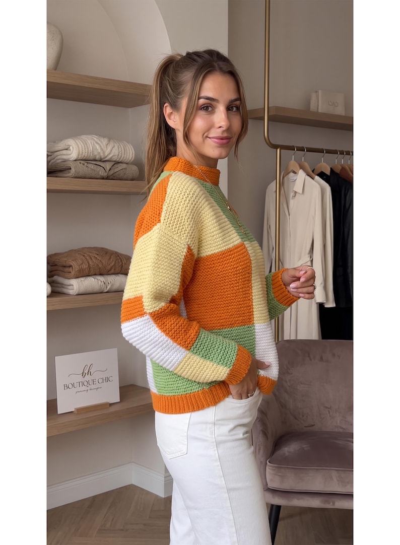 May include: A long-sleeve knit sweater featuring a color-blocked pattern of orange, pale yellow, white, and sage green squares. The sweater has a mock neck collar, ribbed cuffs, and a ribbed hem, paired with plain white pants.