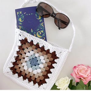 May include: White crochet bag with a brown, beige, and blue granny square design. The bag has a long strap and is shown with a book and sunglasses inside.