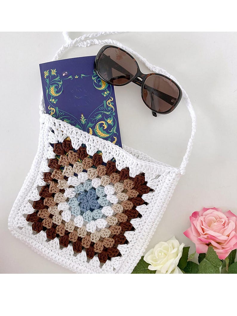 May include: White crochet bag with a brown, beige, and blue granny square design. The bag has a long strap and is shown with a book and sunglasses inside.