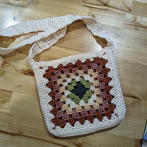 May include: A crocheted bag with a white strap and a colorful granny square design. The bag is made with a cream colored yarn and features shades of brown, green, and beige.