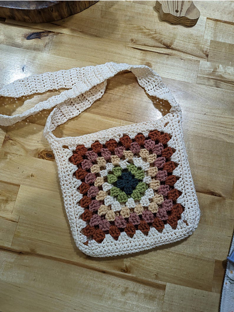 May include: A crocheted bag with a white strap and a colorful granny square design. The bag is made with a cream colored yarn and features shades of brown, green, and beige.