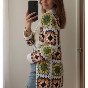 Puede incluir: Un cárdigan blanco de crochet con coloridos patrones cuadrados en tonos azules, naranjas, amarillos y verdes. El suéter de manga larga presenta un diseño clásico de granny square, ideal para un estilo vintage o bohemio.