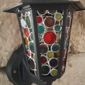 Op de afbeelding: Zwarte buitenlamp voor aan de muur met een glas-in-lood paneel. Het glas heeft een patroon van gekleurde cirkels in rood, groen, geel en wit. De lantaarn is bevestigd aan een getextureerde stenen muur.
