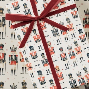 Peut inclure: Un papier cadeau blanc avec un motif répété de soldats casse-noisettes en rouge, bleu et or. Le papier cadeau est noué avec un ruban de velours rouge.