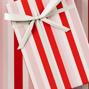 Puede incluir: Una caja de regalo rectangular con un patrón de rayas rojas y blancas, atada con una cinta y un lazo de satén blanco. La caja se coloca sobre un fondo a rayas a juego, creando una presentación festiva y cohesiva.