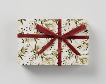 May include: A gift box wrapped in white paper with a floral pattern featuring green leaves and black berries. The box is tied with a red velvet ribbon.