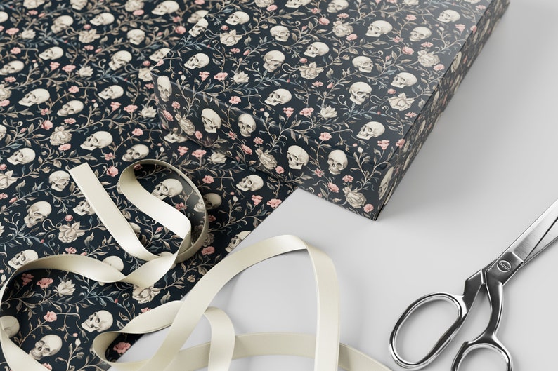 May include: Black and white skull pattern wrapping paper with pink flowers and vines. The paper is wrapped around a box and there is a white ribbon and silver scissors on a grey surface.