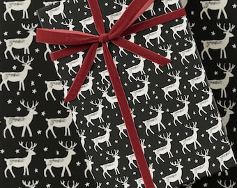 May include: A black gift box with a red velvet ribbon. The box is wrapped in paper with a repeating pattern of white reindeer and stars.