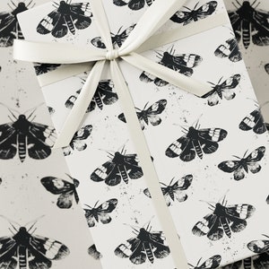 Può includere: Una scatola regalo bianca con un nastro di raso bianco legato in un fiocco. La scatola è avvolta in carta con un motivo a falena bianco e nero.