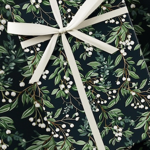 Könnte beinhalten: Eine rechteckige Geschenkbox, eingewickelt in dunkelblaues Papier mit einem sich wiederholenden botanischen Muster aus grünen Blättern und kleinen weißen Beeren. Ein glattes, cremefarbenes Band ist zu einer Schleife auf dem Paket gebunden.