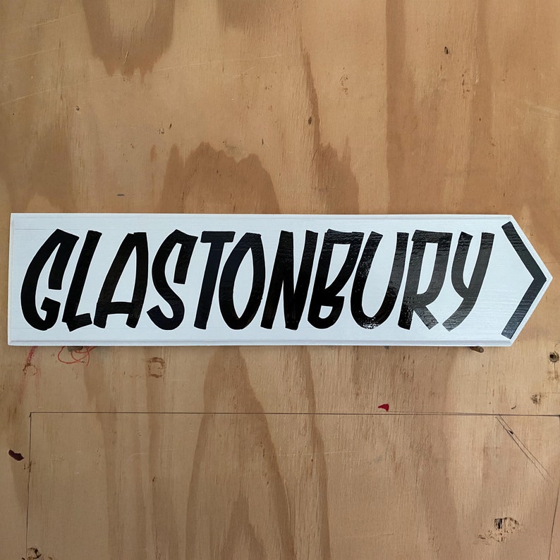 May include: White sign with black lettering pointing to the right that reads "Glastonbury".