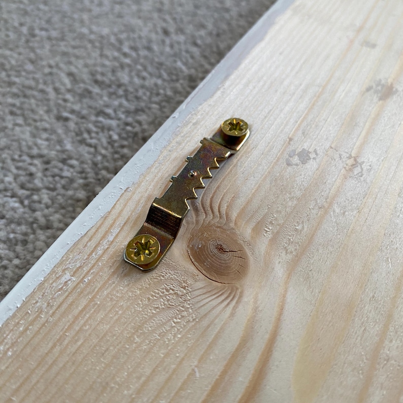 May include: Close-up of a wooden sign with a gold metal hanging hook attached to the back. The hook is secured with two screws.
