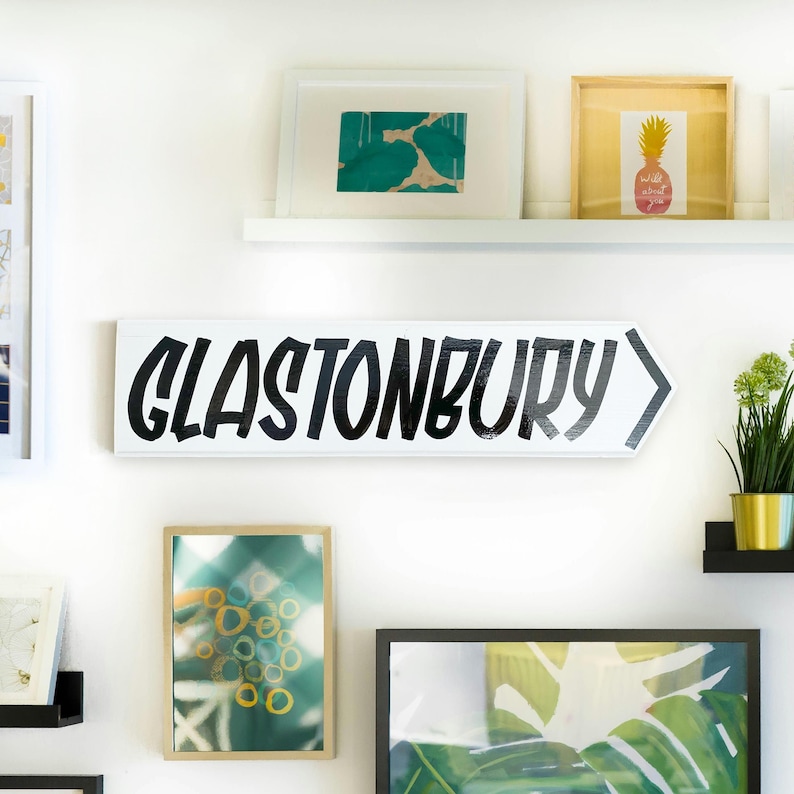 May include: White wooden sign with black painted text that reads "GLASTONBURY" with a black arrow pointing to the right.