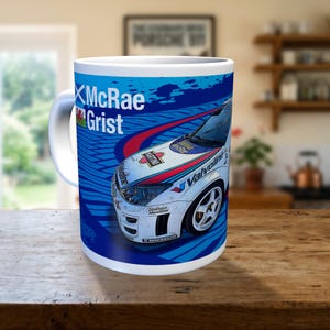 May include: A white ceramic mug featuring a graphic of a white rally car with blue and red racing stripes. The text McRae Grist is printed in white on a blue background, accompanied by a Scottish flag and a Welsh flag icon on the side of the mug.
