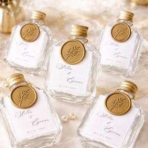 May include: Five clear glass bottles with gold-colored lids and seals. Each bottle has a white label with elegant script and the date 26/12/2025. The seals feature a leaf design. The bottles are arranged on a light-colored surface.