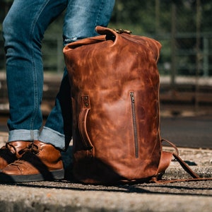 Duffel Bag made of Light Brown Leather, on the ground next to a man who's wearing jeans and brown shoes.