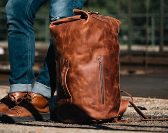 Può includere: Una grande borsa da viaggio in pelle marrone con chiusura a rullo, una tasca frontale verticale con cerniera e una maniglia laterale. La borsa presenta una finitura vissuta ed è realizzata con cuciture visibili lungo i bordi.
