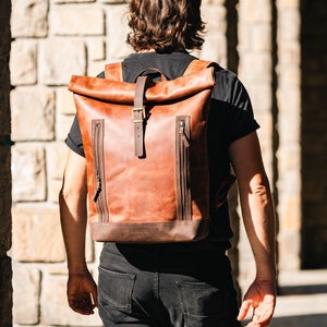 Pode incluir: Uma mochila de couro castanho-claro com fecho de enrolar, uma correia vertical central com fivela de metal, dois bolsos laterais com fecho e puxadores de couro, e uma base reforçada em castanho-escuro, usada sobre t-shirt e calças pretas.