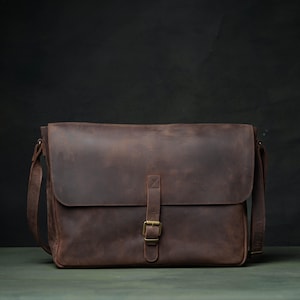 May include: A dark brown leather messenger bag featuring a large front flap, a single vertical strap with a brass-toned metal buckle closure, and an adjustable shoulder strap. The bag has a smooth, matte finish and a rectangular silhouette.