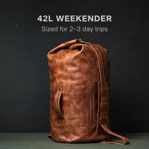Puede incluir: Una bolsa de viaje weekender de 42L en cuero marr&oacute;n con cierre enrollable, un asa lateral y un bolsillo vertical con cremallera. El texto 42L WEEKENDER Sized for 2-3 day trips es visible sobre la bolsa, que presenta costuras y correas reforzadas.