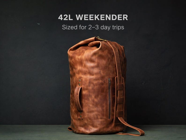 Puede incluir: Una bolsa de viaje weekender de 42L en cuero marr&oacute;n con cierre enrollable, un asa lateral y un bolsillo vertical con cremallera. El texto 42L WEEKENDER Sized for 2-3 day trips es visible sobre la bolsa, que presenta costuras y correas reforzadas.