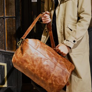 Puede incluir: Una bolsa de viaje de cuero color canela con una textura suave y desgastada, que cuenta con una correa larga ajustable para el hombro con herrajes de metal color lat&oacute;n, un asa superior y una forma rectangular con bordes redondeados.