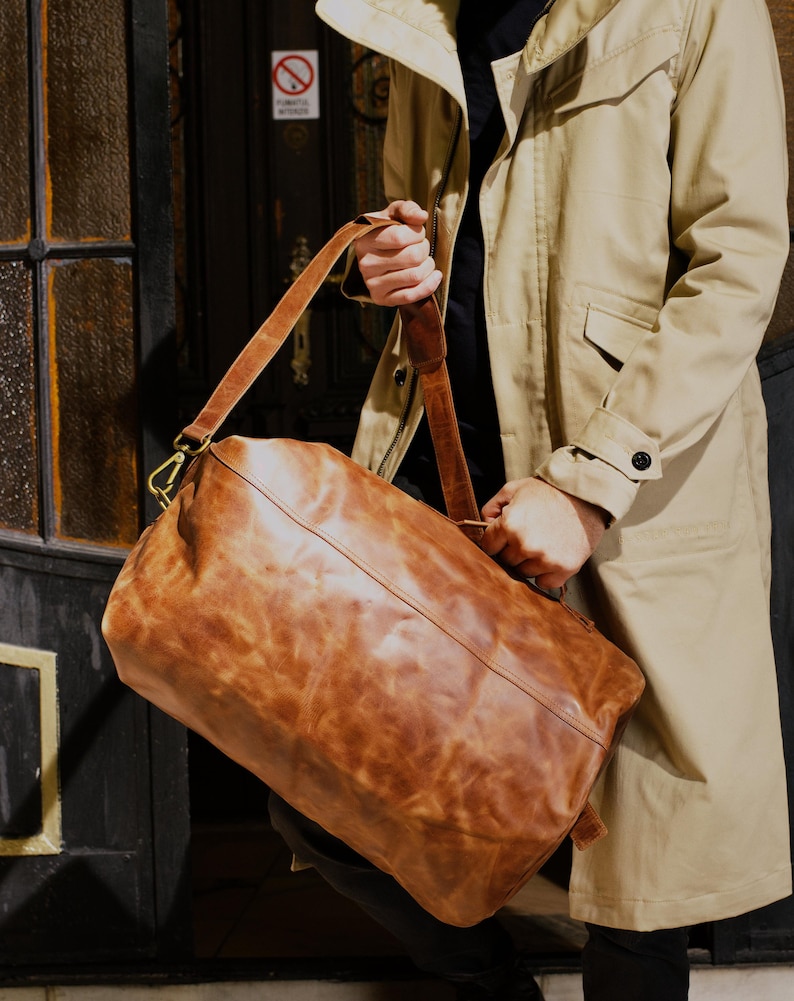 Puede incluir: Una bolsa de viaje de cuero color canela con una textura suave y desgastada, que cuenta con una correa larga ajustable para el hombro con herrajes de metal color lat&oacute;n, un asa superior y una forma rectangular con bordes redondeados.