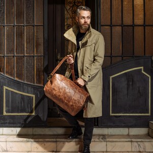 Puede incluir: Una gabardina color canela sobre una camisa negra y pantalones oscuros, acompa&ntilde;ada de una bolsa de viaje cil&iacute;ndrica de cuero marr&oacute;n con herrajes de lat&oacute;n, un asa superior resistente y una correa de hombro ajustable para llevar objetos.