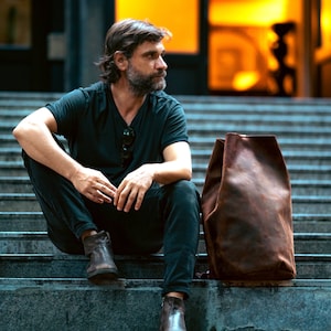 A cylindrical duffel bag sitting next to a stylish man with beard on stairs. The bag is made of leather and it's brown.