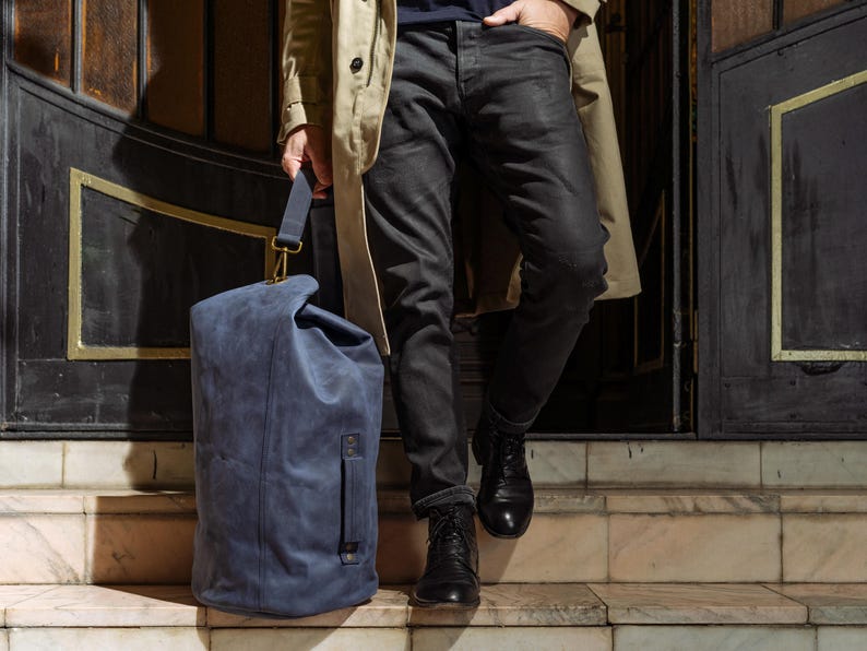Puede incluir: Una bolsa de viaje azul con cierre enrollable, un asa lateral y una correa de hombro extra&iacute;ble con herrajes de lat&oacute;n. La bolsa est&aacute; hecha de un material mate y texturizado, y presenta una costura vertical en el panel frontal.