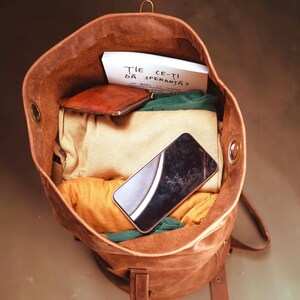 Puede incluir: Una mochila de cuero marr&oacute;n llena de ropa doblada, una peque&ntilde;a cartera de cuero marr&oacute;n, un tel&eacute;fono inteligente negro y una tarjeta blanca con el texto "ȚIE CE-ȚI DĂ SPERANȚĂ?" impreso en tinta negra. La mochila tiene ojales met&aacute;licos laterales.