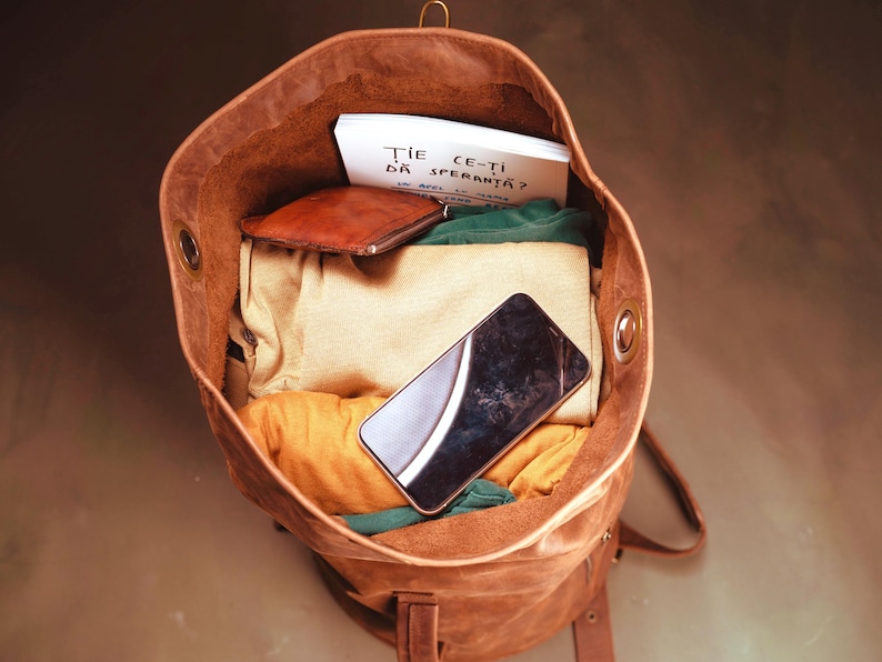 Puede incluir: Una mochila de cuero marr&oacute;n llena de ropa doblada, una peque&ntilde;a cartera de cuero marr&oacute;n, un tel&eacute;fono inteligente negro y una tarjeta blanca con el texto "ȚIE CE-ȚI DĂ SPERANȚĂ?" impreso en tinta negra. La mochila tiene ojales met&aacute;licos laterales.
