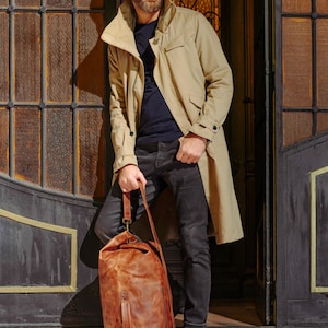 Puede incluir: Una gabardina color canela sobre una camiseta azul oscuro y vaqueros gris oscuro, combinada con botas negras. Un bolso de viaje grande de cuero marr&oacute;n desgastado, con asa superior y correa para el hombro, se sit&uacute;a frente a las piernas.