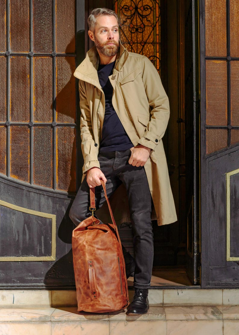 Puede incluir: Una gabardina color canela sobre una camiseta azul oscuro y vaqueros gris oscuro, combinada con botas negras. Un bolso de viaje grande de cuero marr&oacute;n desgastado, con asa superior y correa para el hombro, se sit&uacute;a frente a las piernas.
