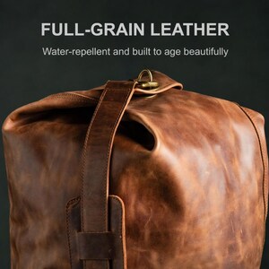 Puede incluir: Una bolsa de cuero de grano completo en color marr&oacute;n con cierre enrollable, una correa vertical de cuero y un anillo met&aacute;lico en tono lat&oacute;n. El texto FULL-GRAIN LEATHER y Water-repellent and built to age beautifully aparece arriba.