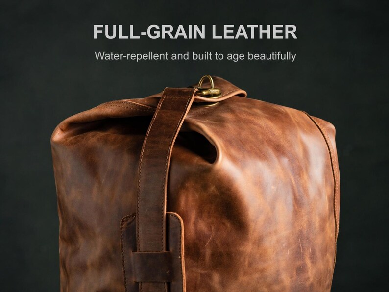 Puede incluir: Una bolsa de cuero de grano completo en color marr&oacute;n con cierre enrollable, una correa vertical de cuero y un anillo met&aacute;lico en tono lat&oacute;n. El texto FULL-GRAIN LEATHER y Water-repellent and built to age beautifully aparece arriba.