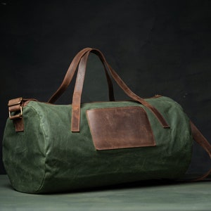 May include: Green duffel bag with brown leather accents. Features include a brown leather patch and handles, plus a detachable shoulder strap. The bag is made of a durable, water-resistant material, perfect for travel or the gym.