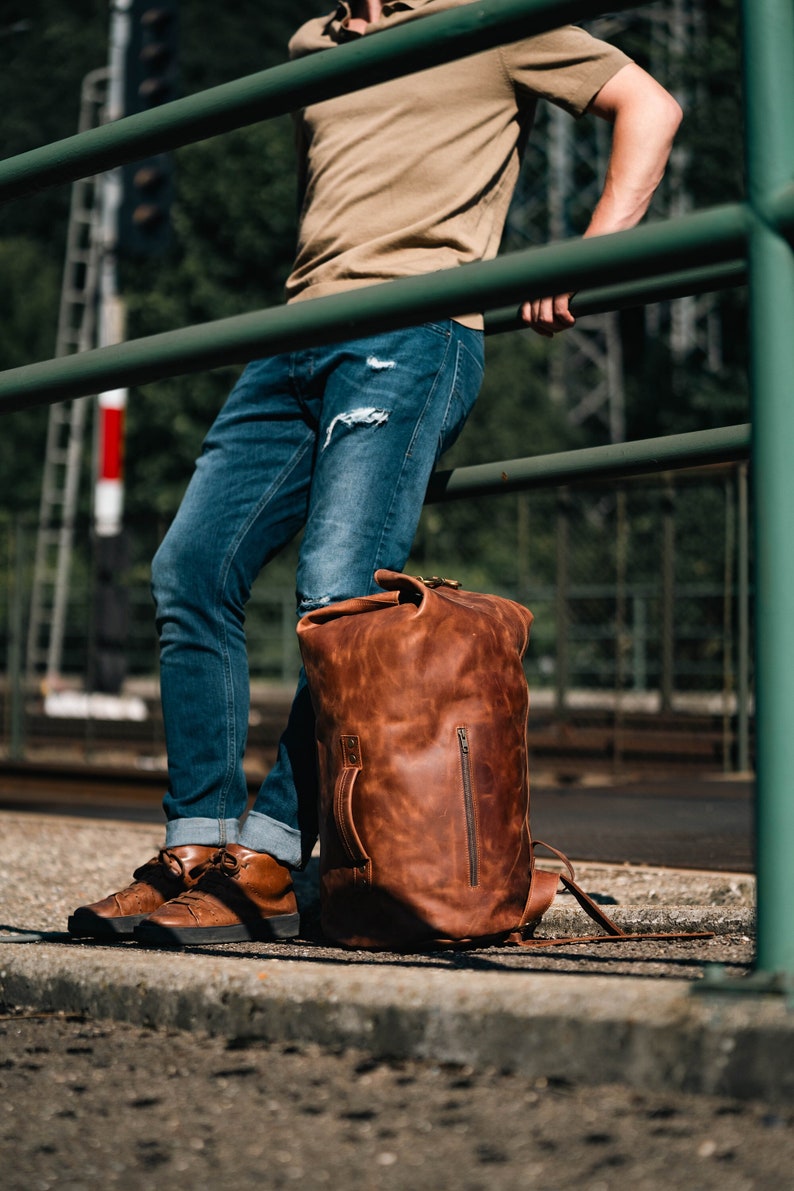 Duffel Bag made of Light Brown Leather, on the ground next to a man who's wearing jeans and brown shoes.