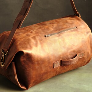 Puede incluir: Una bolsa de viaje de cuero marr&oacute;n con forma cil&iacute;ndrica, que cuenta con un asa lateral asegurada con remaches met&aacute;licos, un bolsillo exterior con cremallera y una correa de hombro extra&iacute;ble con ganchos y herrajes de lat&oacute;n.