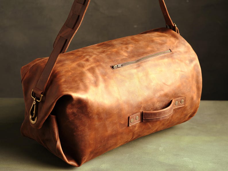 Puede incluir: Una bolsa de viaje de cuero marr&oacute;n con forma cil&iacute;ndrica, que cuenta con un asa lateral asegurada con remaches met&aacute;licos, un bolsillo exterior con cremallera y una correa de hombro extra&iacute;ble con ganchos y herrajes de lat&oacute;n.