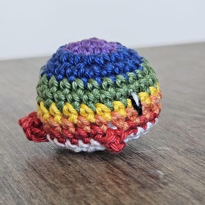 Könnte beinhalten: Ein gehäkelter Regenbogenball mit einem weißen Gesicht und schwarzen Augen. Der Ball ist aus Garn gefertigt und hat eine strukturierte Oberfläche.