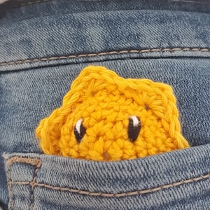 Könnte beinhalten: Ein gelber Häkelstern mit einem lächelnden Gesicht steckt in einer blauen Jeans-Tasche.
